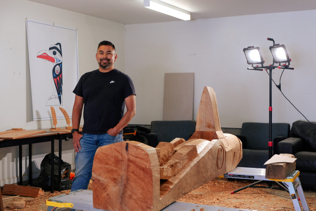 Bear Horne Kevin Mccarthy standing in a workshop with a large wooden carving.