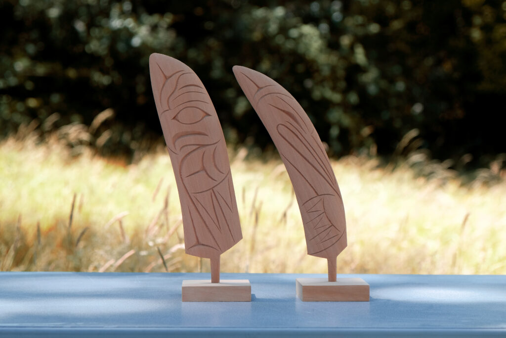 Two wooden carvings of stylized feathers with intricate etchings, displayed on stands against a blurred outdoor background.